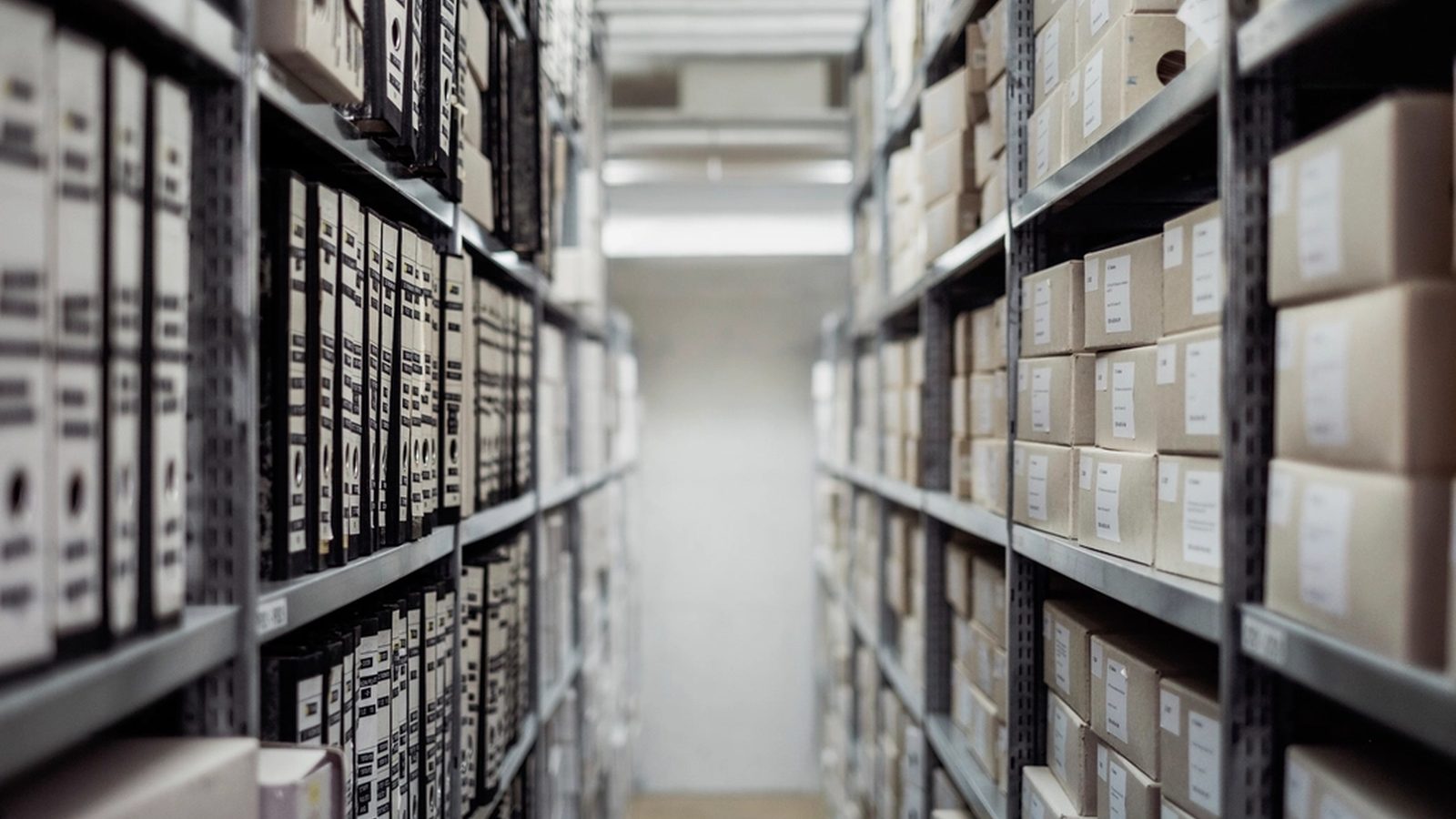 View down an archive aisle: shelves of ring binders on the left, shelves of labeled archive boxes on the right, bright light at the end of the aisle.