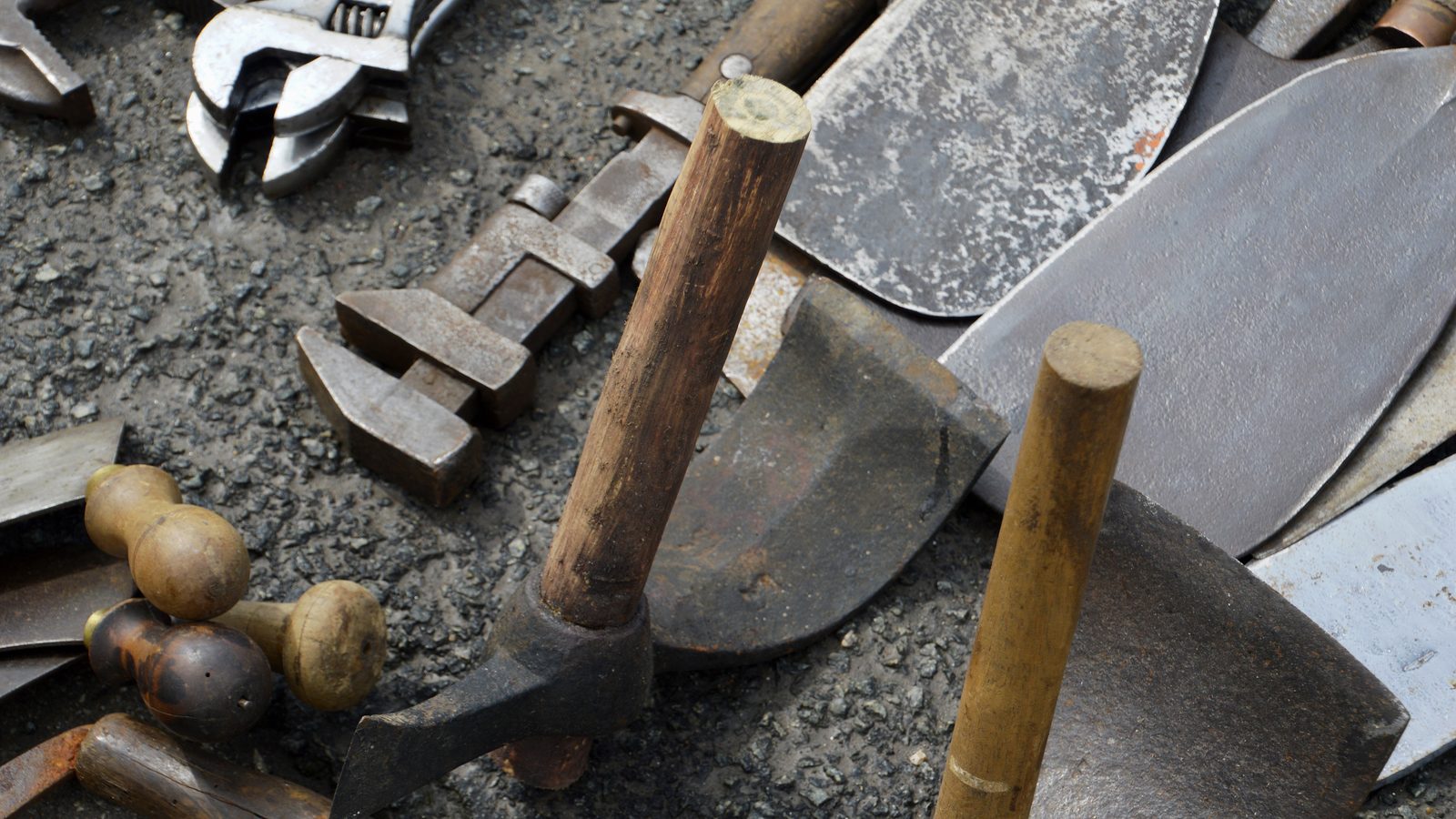 Used hand tools on a gravel surface: hammers with wooden handles, adjustable wrenches, and shovel blades.