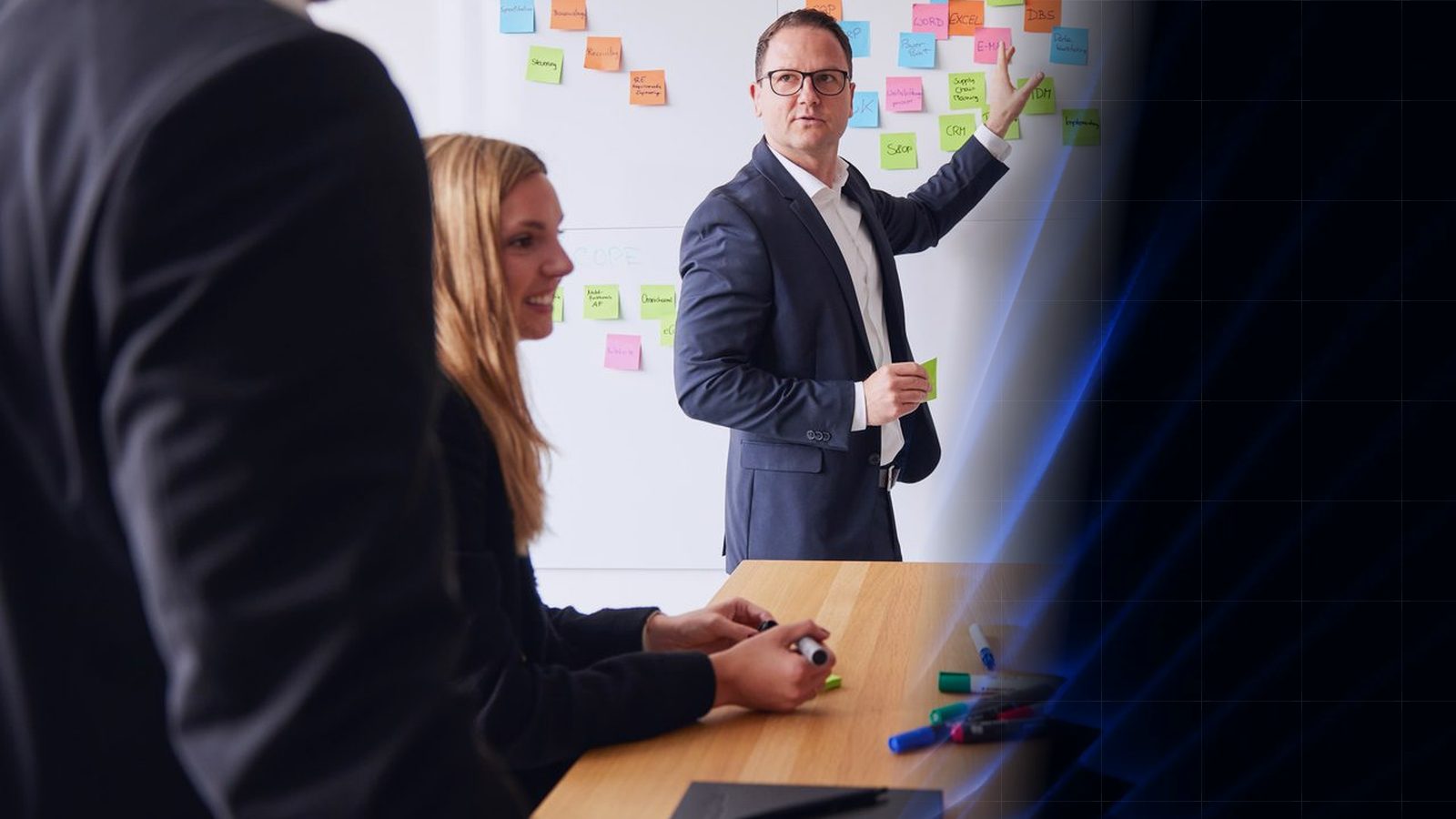 Workshop at ReqPOOL: a consultant points at a wall of colored sticky notes while a colleague takes notes at the table; on the right, the image blends into the blue shape pattern of the ReqPOOL website.