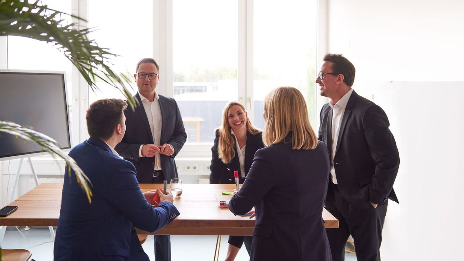 Five ReqPOOL consultants in discussion around a tall wooden table in a bright meeting room with a screen and an indoor palm.