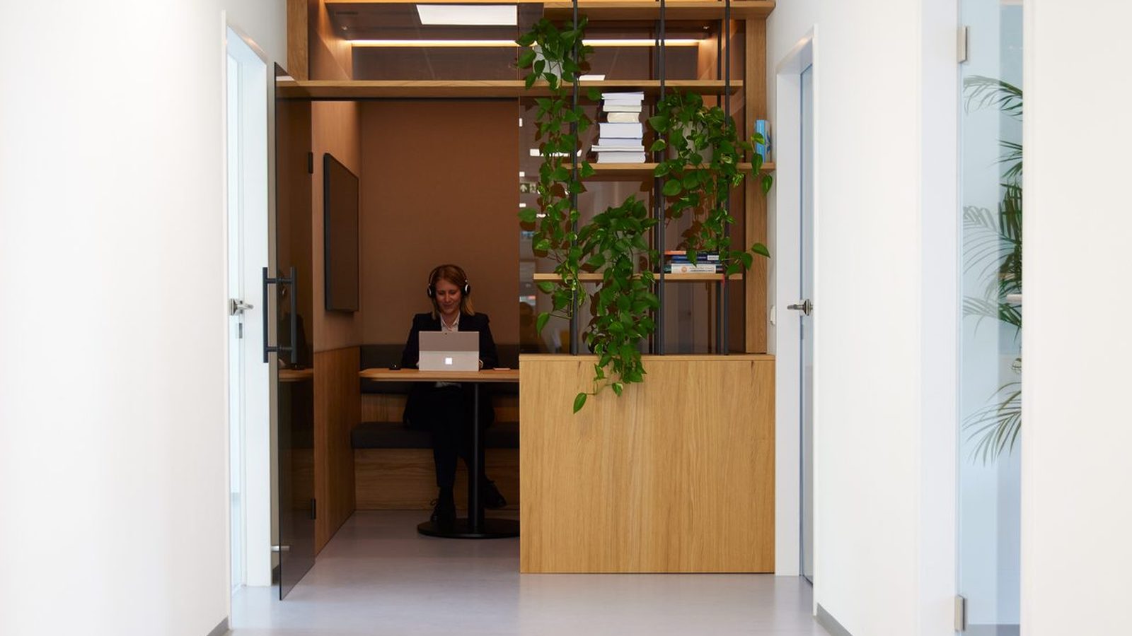 View along a bright office corridor at ReqPOOL into a wooden meeting booth with plants, where an employee is working on a laptop.