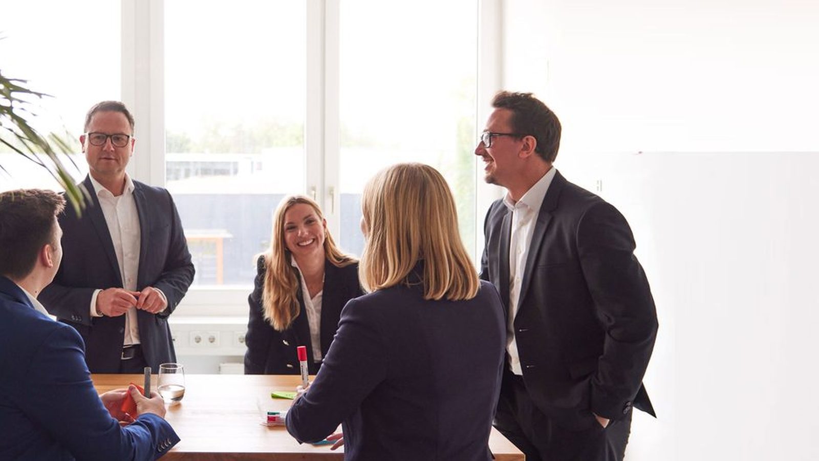 Meeting at ReqPOOL: five consultants stand and sit around a wooden table in a bright office with a large screen.