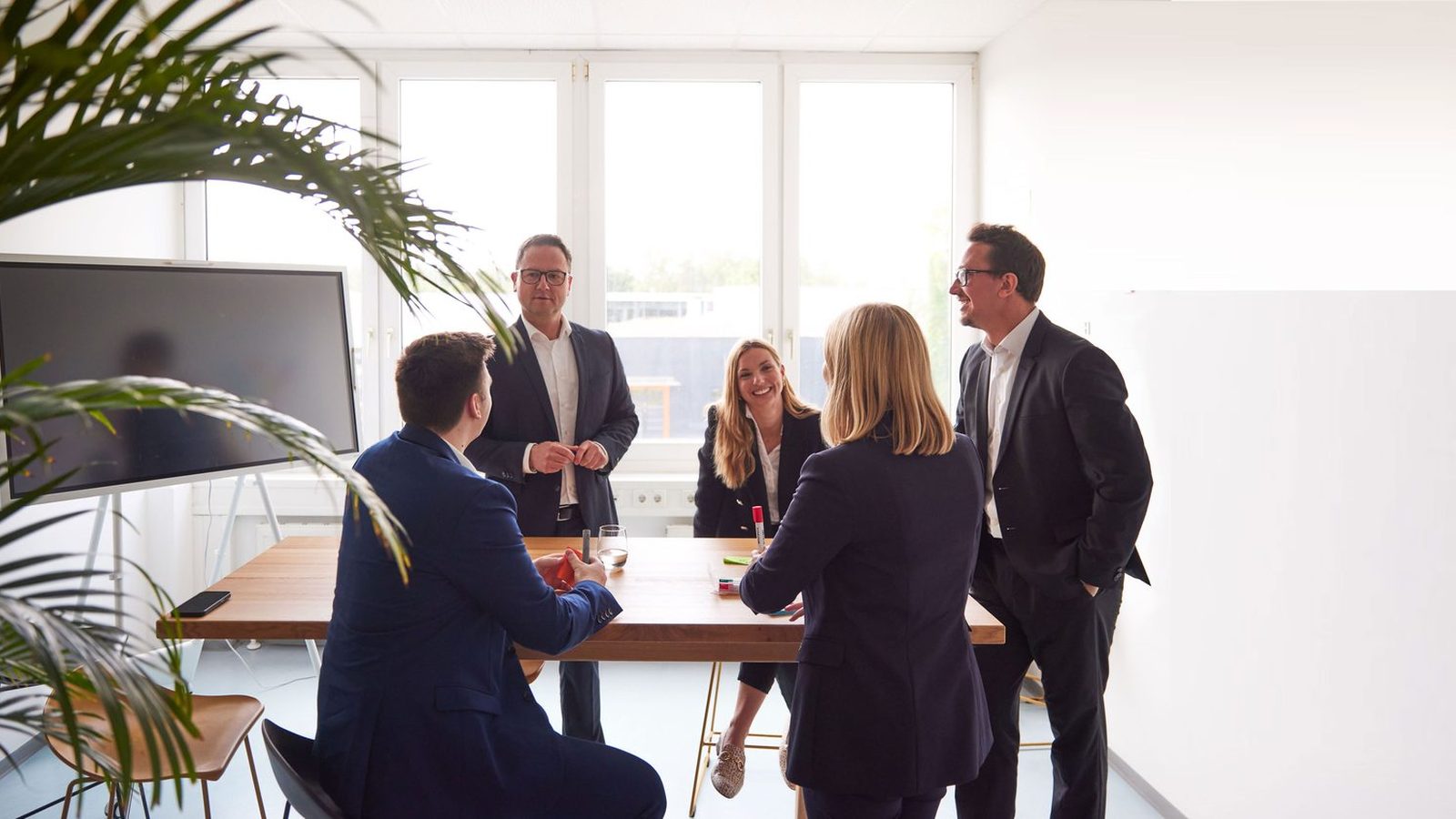 Five ReqPOOL consultants discuss a decision at a standing table in a bright meeting room.
