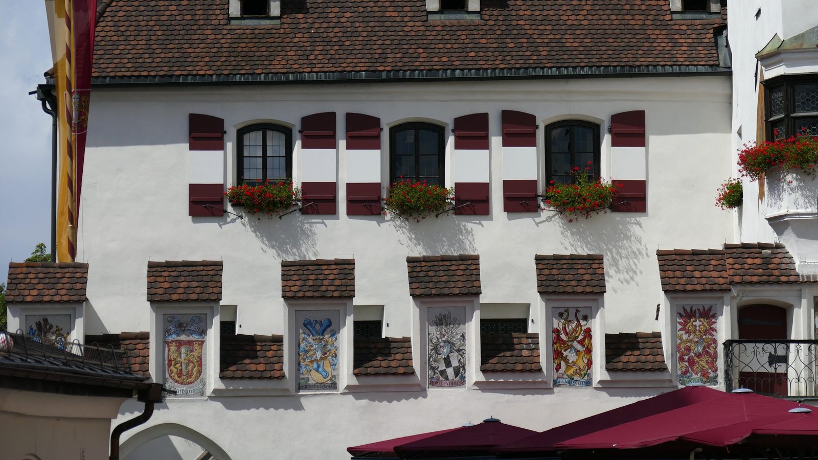 Facade of the town hall on the Upper Town Square in Hall in Tirol with dark red shutters, flower boxes, and historic coats of arms painted on the wall.