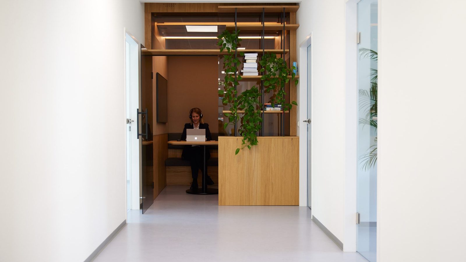 View down a bright ReqPOOL office corridor toward a consultant wearing headphones and working on a laptop in a wood-paneled alcove.