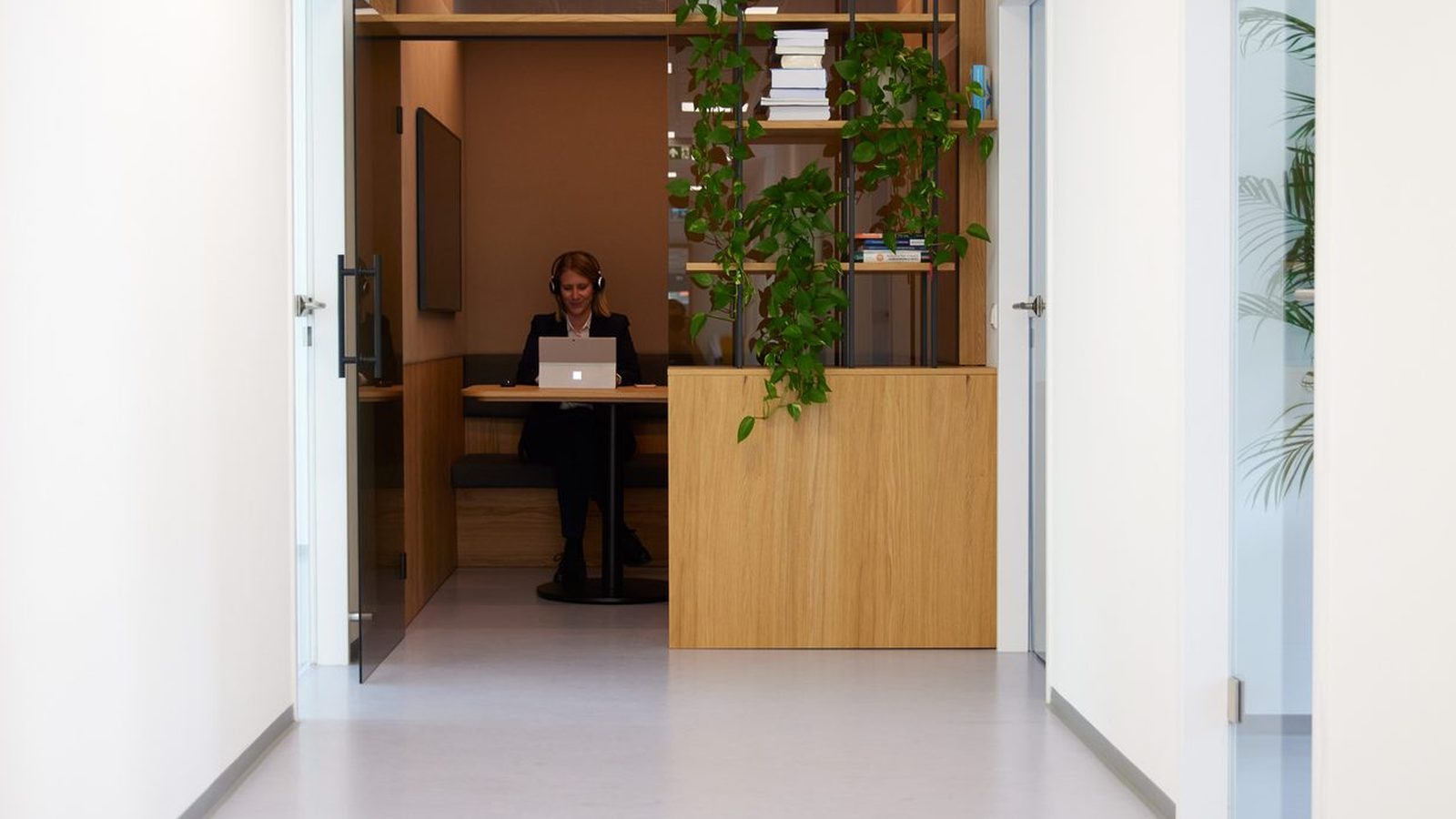 Bright office corridor at ReqPOOL; at its end a consultant wearing headphones works on a laptop in a wooden alcove, next to a shelf with plants and books.