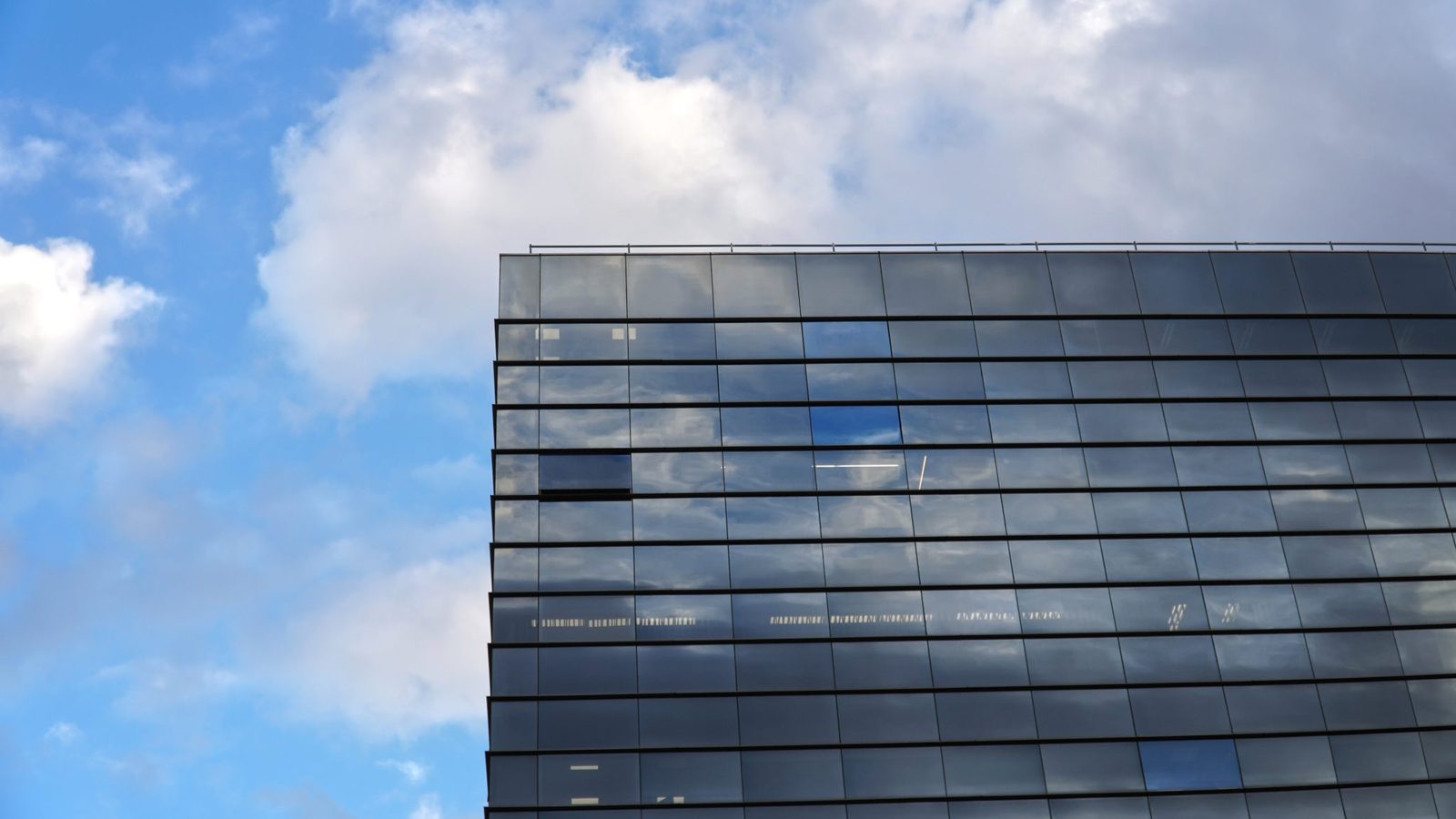 Glass facade of a modern office building photographed from below, its windows reflecting a blue sky with white clouds.