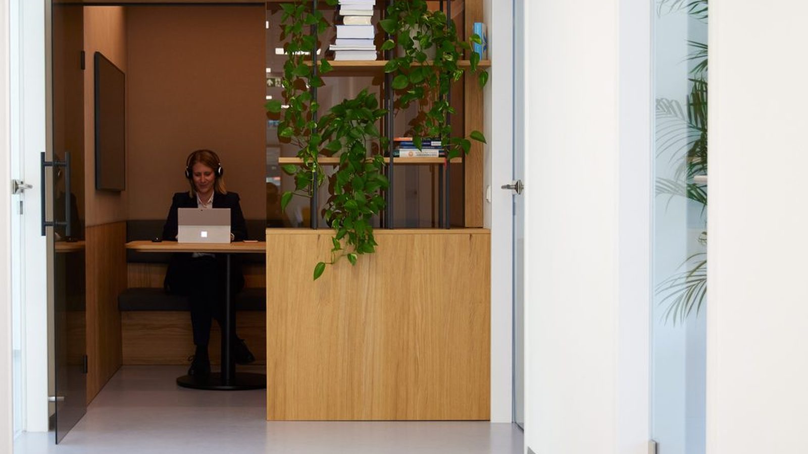 Bright office corridor at ReqPOOL, at the end of which a consultant wearing headphones works on a laptop in a wooden alcove, next to a shelf with plants and books.