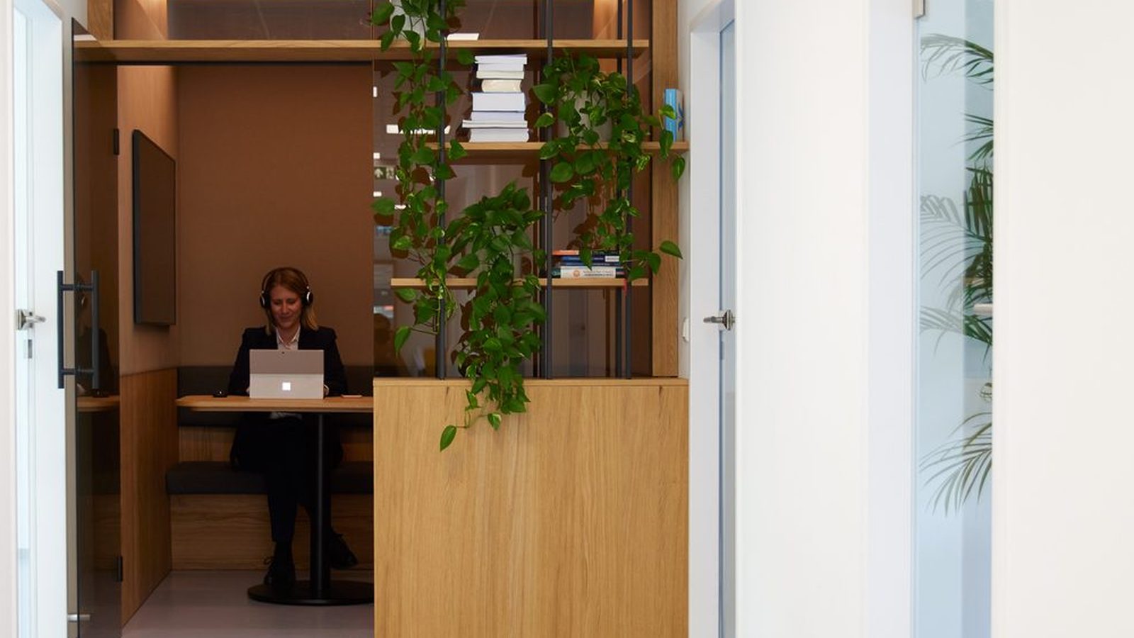Bright corridor in a ReqPOOL office: through an open glass door, a consultant wearing a headset works on a laptop in a wooden alcove, next to a shelf with plants.
