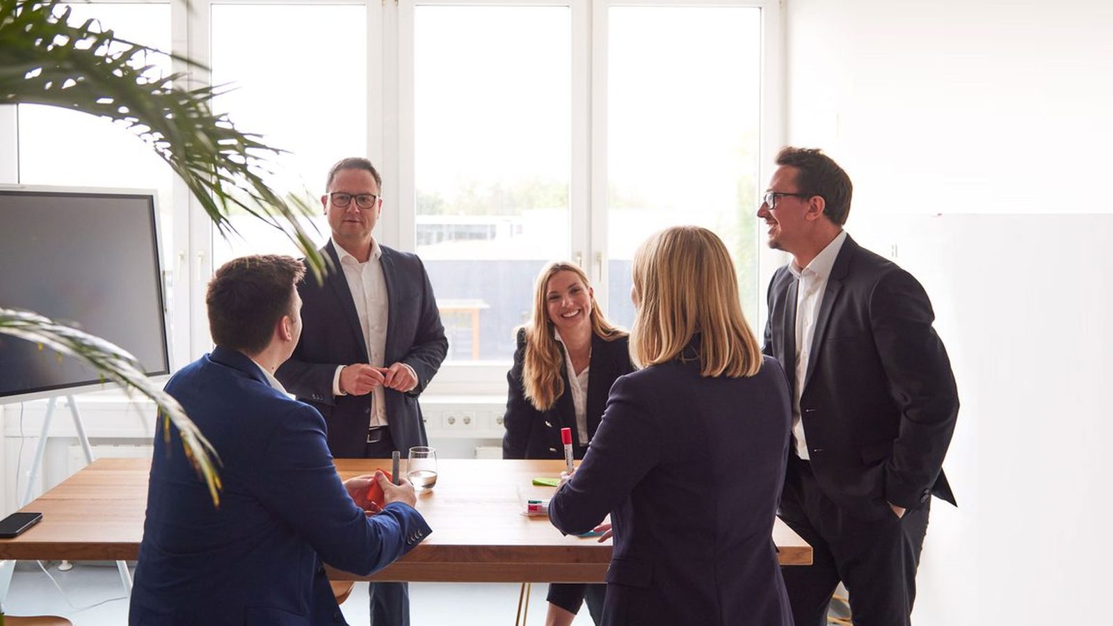 Five ReqPOOL consultants in discussion around a tall wooden table in a bright office with a screen and a houseplant.