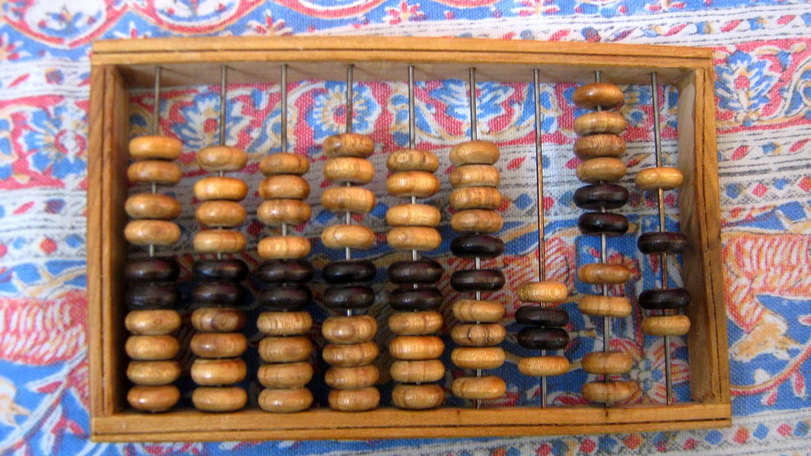 A wooden abacus with light and dark wooden beads lies on a blue and red patterned cloth.