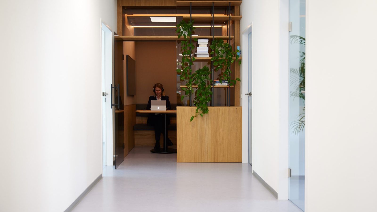 Bright office corridor at ReqPOOL; at its end a consultant wearing headphones works on a laptop in a wood-paneled alcove next to a plant-covered shelf.