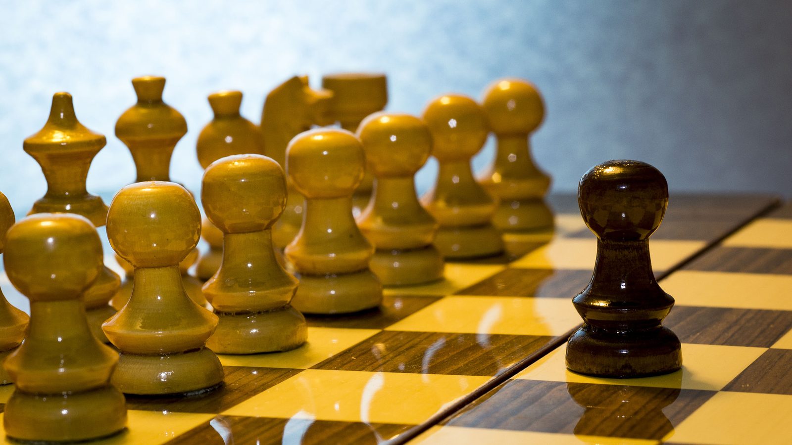 Close-up of a wooden chessboard: a single dark pawn faces a row of light pieces.