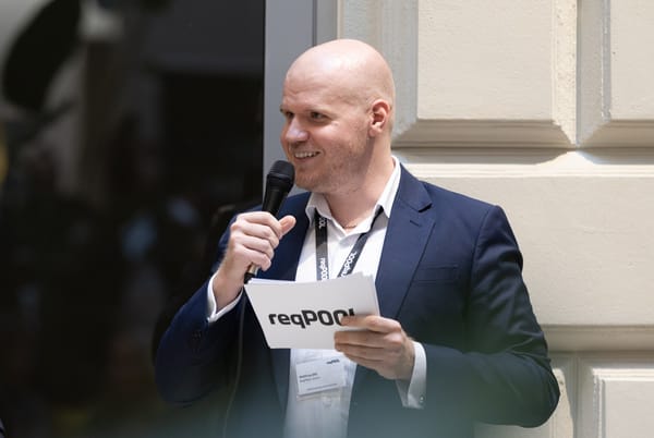 Smiling speaker with a microphone and a ReqPOOL cue card in front of a light facade