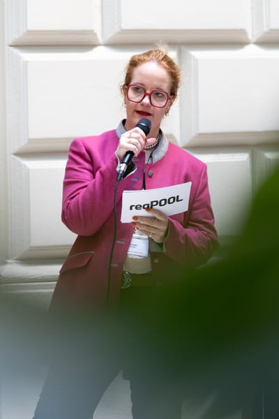 Speaker in a pink blazer talking into a microphone in front of a light facade