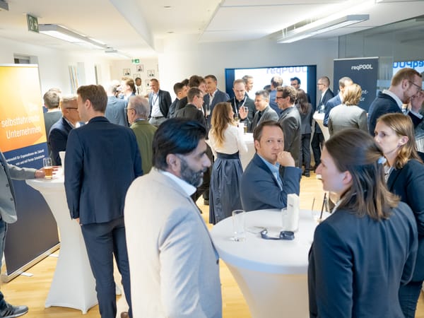 Guests talking at standing tables in the Vienna office, with a banner for the book “The Self-Driving Company” on the left