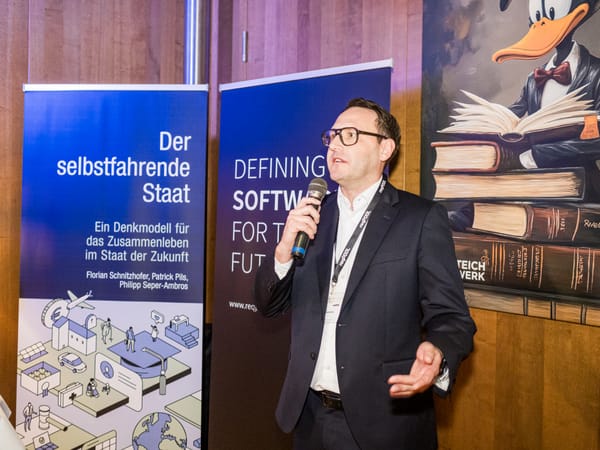 Redner mit Mikrofon vor dem Banner „Der selbstfahrende Staat“