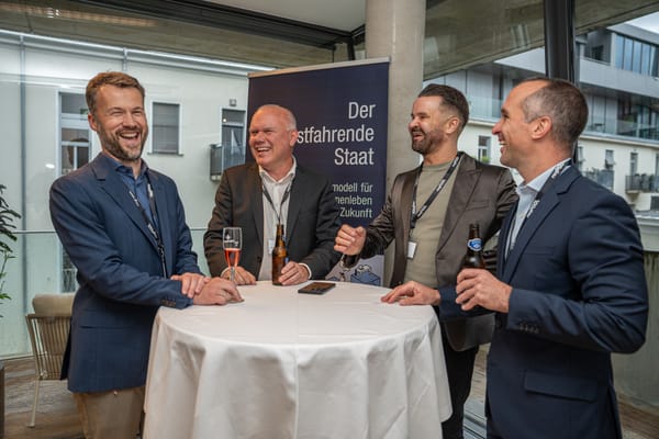 Four men laughing at a standing table on the terrace, with a banner on “The Self-Driving State” behind them