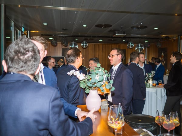 Gäste im Gespräch an einer Bar im Veranstaltungsraum, im Vordergrund Cocktailgläser und eine Vase